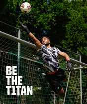 A soccer player in a black and white patterned jersey and black gloves leaps to catch a soccer ball in front of a goal net, with trees in the background and the text 'BE THE TITAN' on the left.